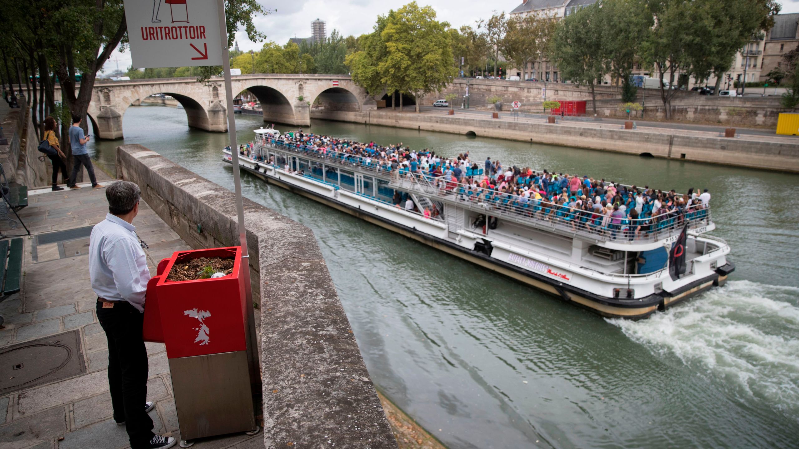 A man stands at a 'uritrottoir' public urinal on August 13, 2018, on the Saint-Louis island in Paris, as a 'bateau mouche' tourist barge cruises past. (Credit: THOMAS SAMSON/AFP/Getty Images)