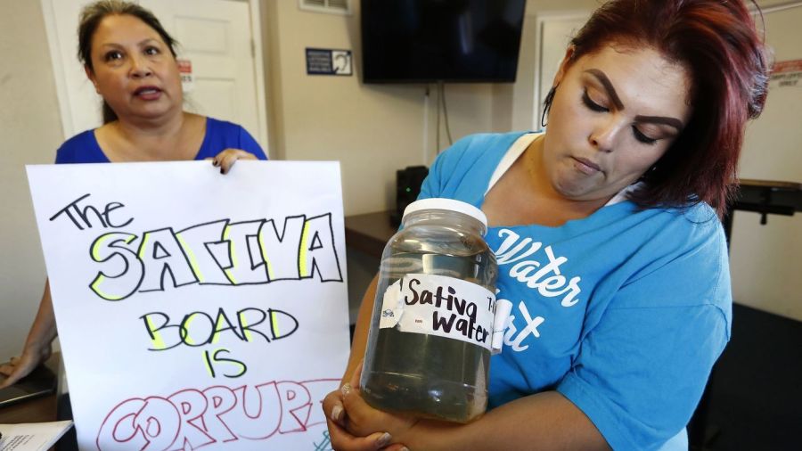 An undated photo shows Compton resident Genoveva Camargo, right, holding water from her tap while activist Maria Estrada displays a sign that reads: "The Sativa Board is corrupt." (Credit: Mel Melcon / Los Angeles Times)