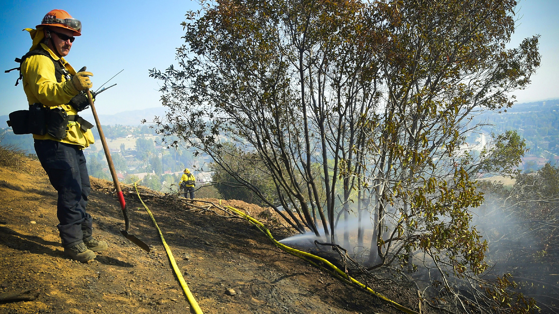 A firefighter overlooks the scene standing beside a a punctured hose spraying out water on a hillside overlooking homes in West Hills, near Malibu, California on November 11, 2018, as the battle to control the Woolsey Fire continues. (Credit: FREDERIC J. BROWN/AFP/Getty Images)