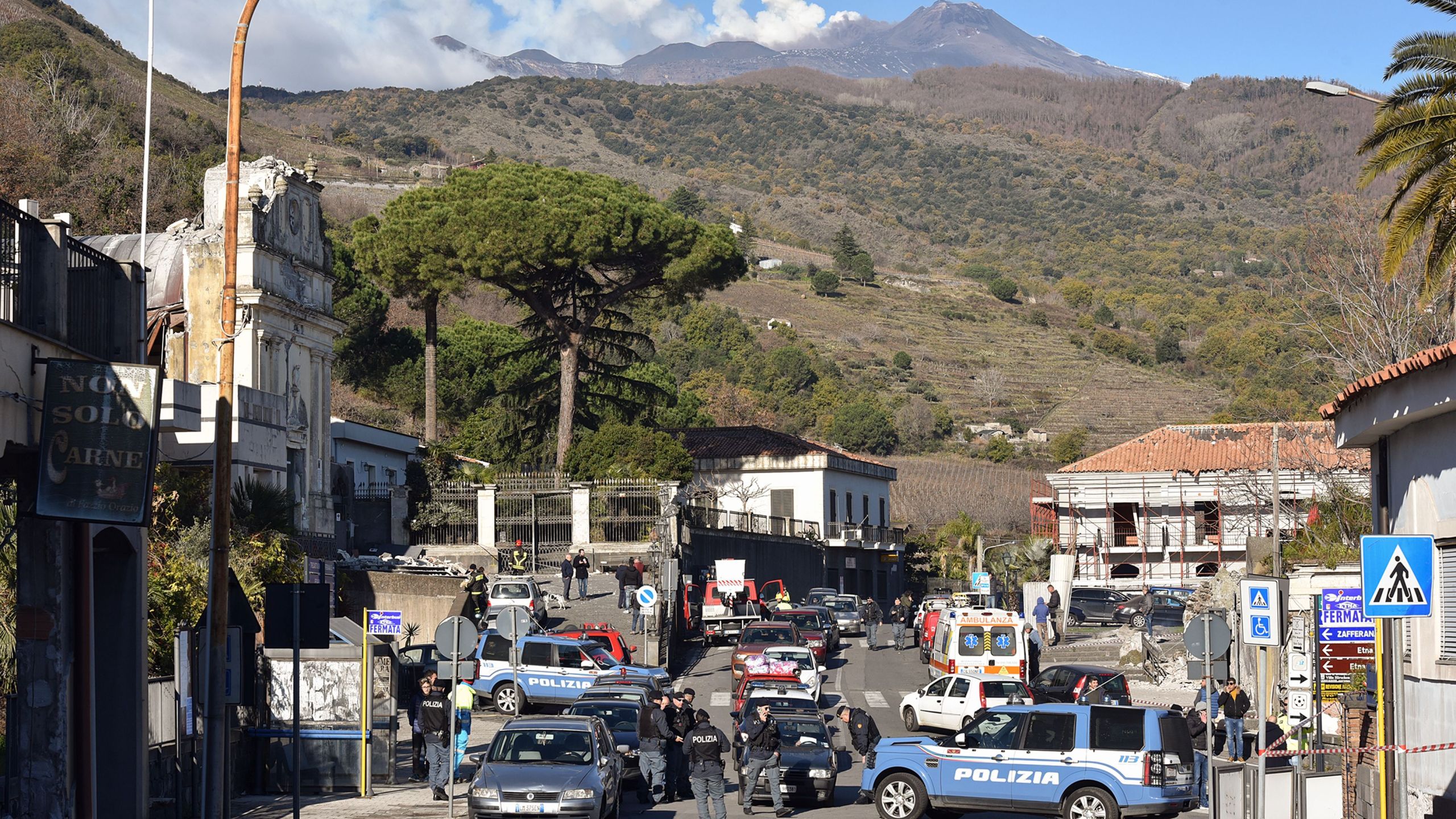 A general view shows police officers and an ambulance outside the damaged church of Parrocchia Maria Ss.Del Rosario (left), with smoke rising from Mount Etana in Zafferana Etnea near Catania on Dec. 26, 2018. (Credit: Giovanni Isolino/AFP/Getty Images)