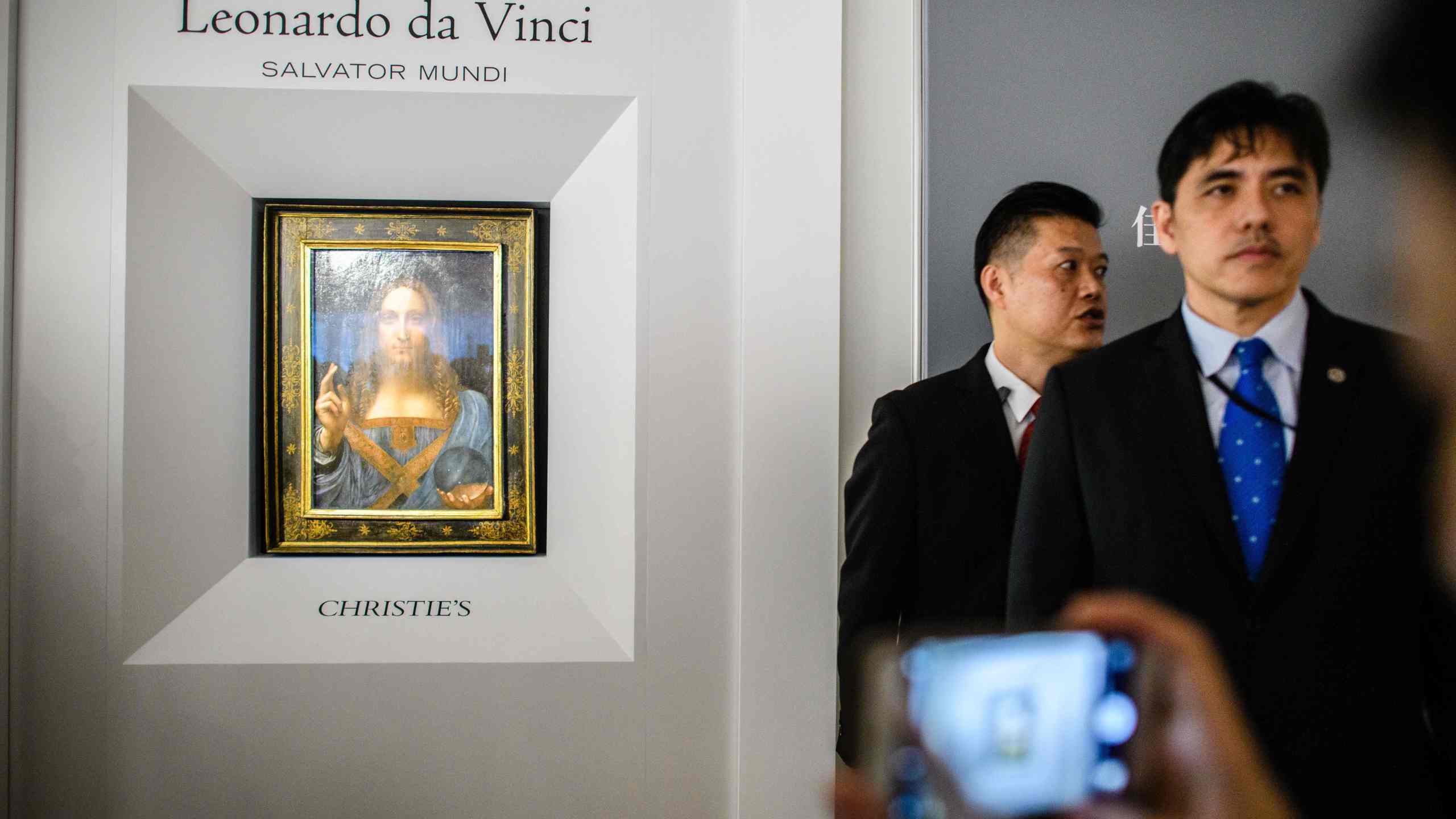 This picture taken on October 13, 2017, shows a man identified by local Hong Kong media as former CIA agent Jerry Chun Shing Lee standing in front of a member of security at the unveiling of Leonardo da Vinci's 'Salvator Mundi' painting at the Christie's showroom in Hong Kong. (Credit: Anthony Wallace/AFP via Getty Images)