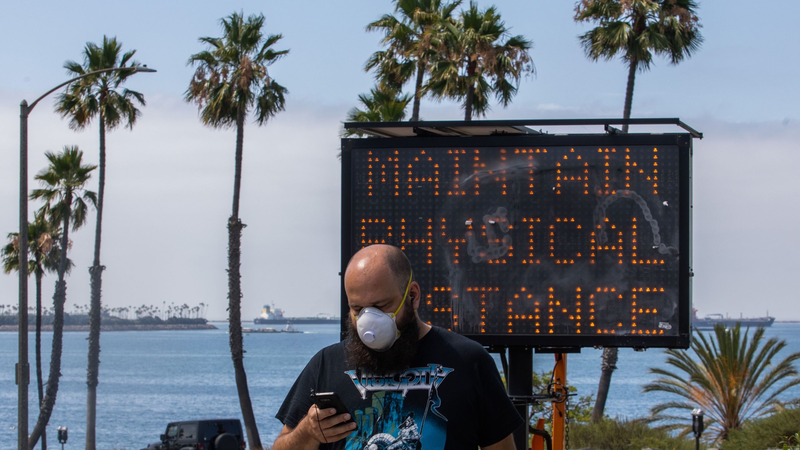 A man wearing a facemask checks his phone near a sign encouraging people to maintain social distancing, while he walks on the beach in Long Beach on July 14, 2020. (APU GOMES/AFP via Getty Images)