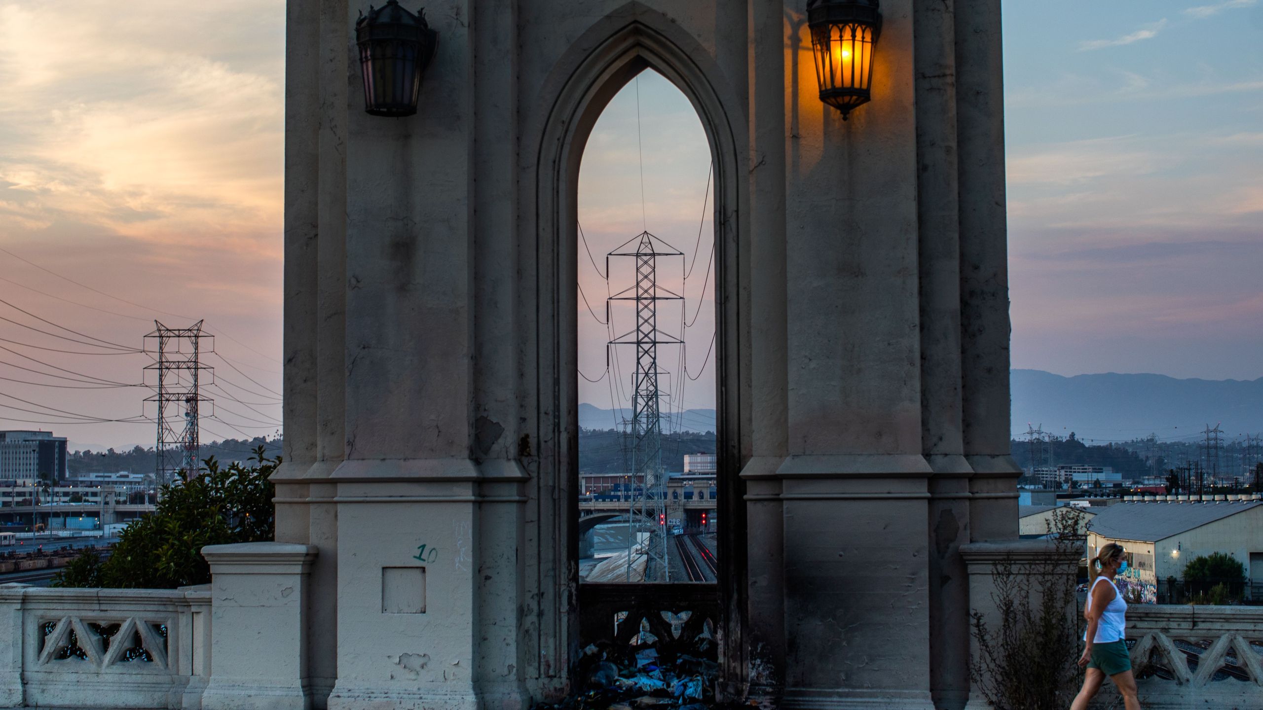 A woman wearing a face mask walks on the Fourth Street Bridge in Los Angeles with high tension towers in the background on Aug. 16, 2020, two days after California on Aug. 14 ordered rolling power outages for the first time since 2001. (Apu Gomes / AFP / Getty Images)