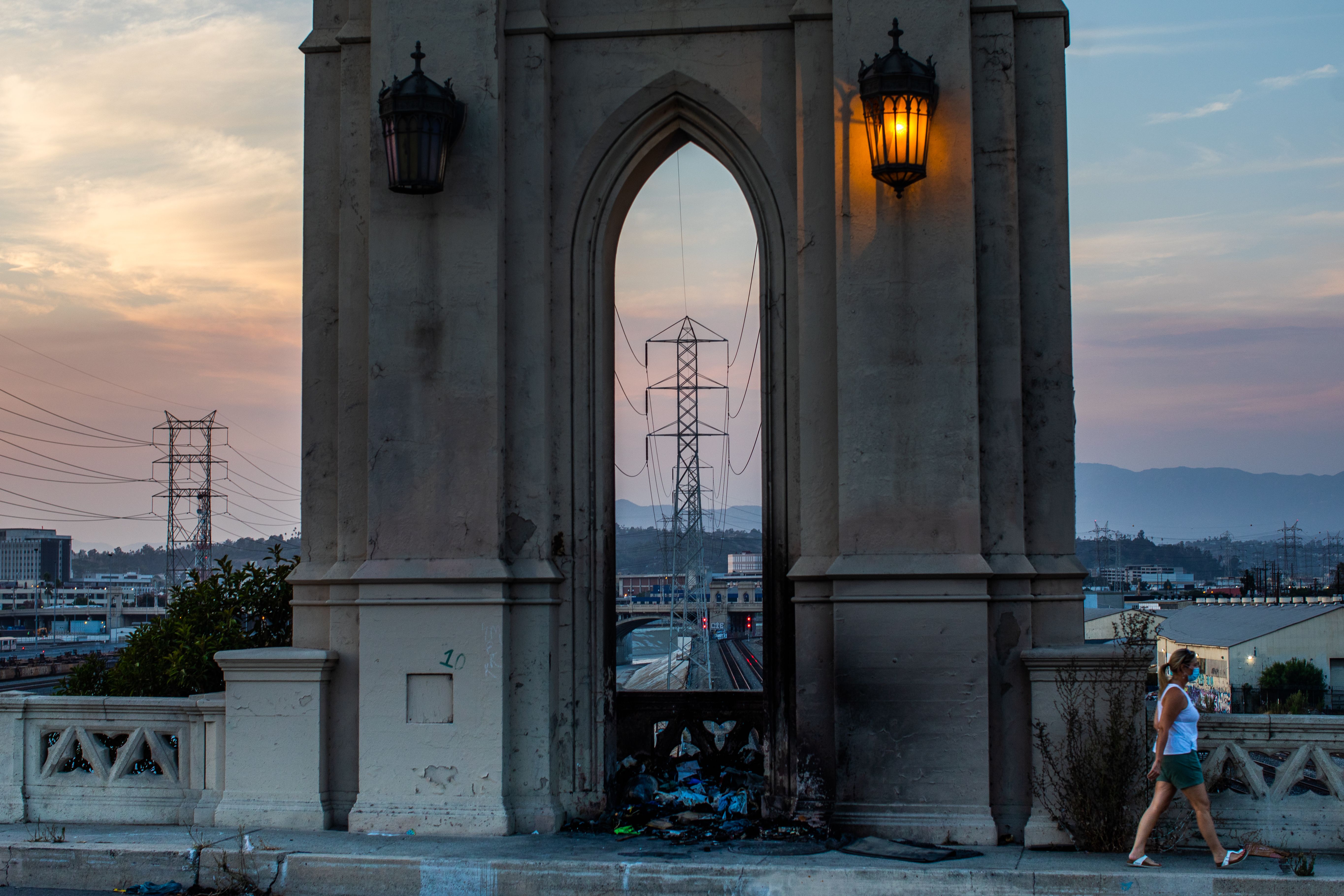 A woman wearing a face mask walks on the Fourth Street Bridge in Los Angeles with high tension towers in the background on Aug. 16, 2020, two days after California on Aug. 14 ordered rolling power outages for the first time since 2001. (Apu Gomes / AFP / Getty Images)