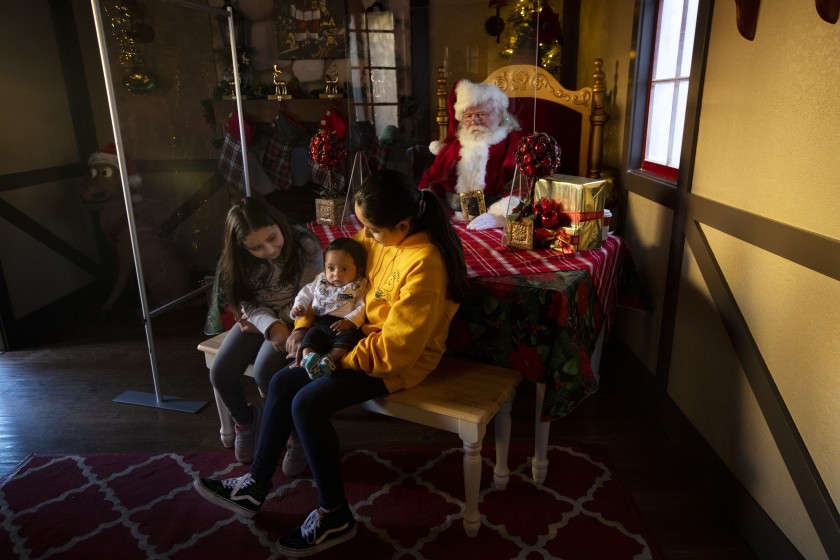 Catherine Mejia, left, Damian Rocha and Lizzette Mejia have their photo taken on Nov. 28, 2020 at the Citadel Outlets with Santa Claus, played by Ray Hamlett. Santa’s new setup included plexiglass shields.(Francine Orr / Los Angeles Times)