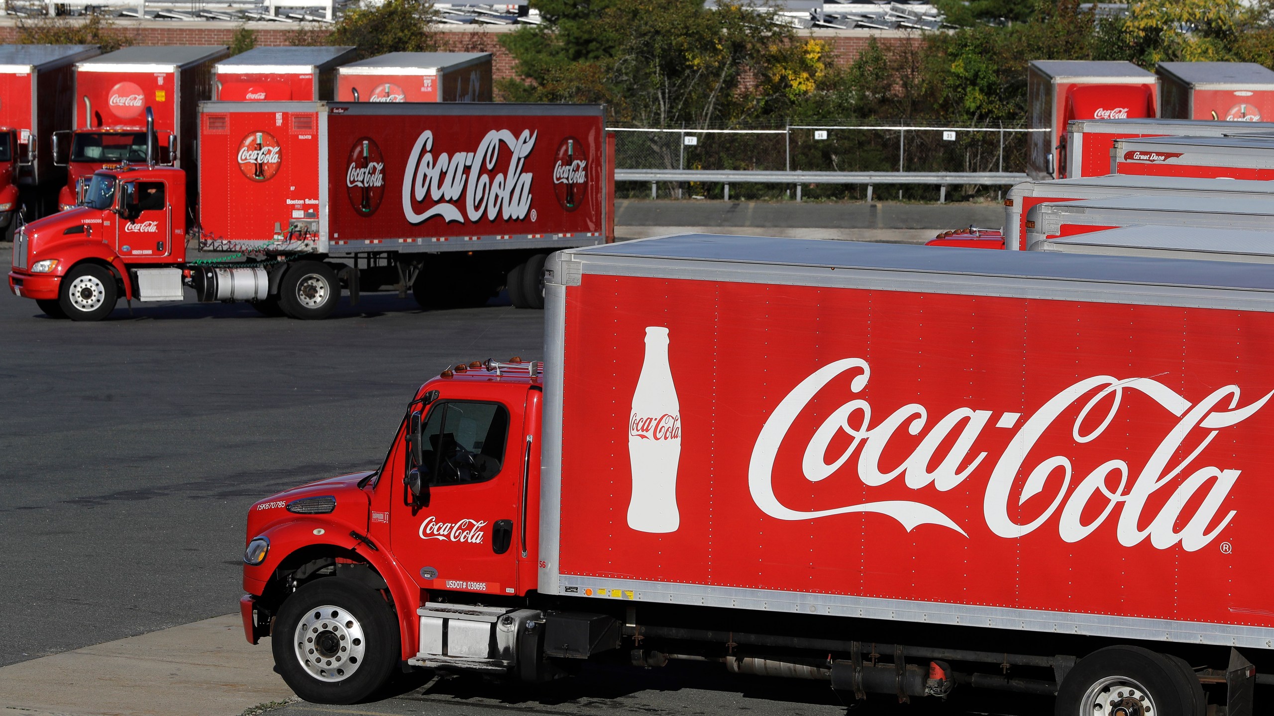 In this Oct. 14, 2019, photo a truck with the Coca-Cola logo, behind left, maneuvers in a parking lot at a bottling plant in Needham, Mass. (AP Photo/Steven Senne, File)