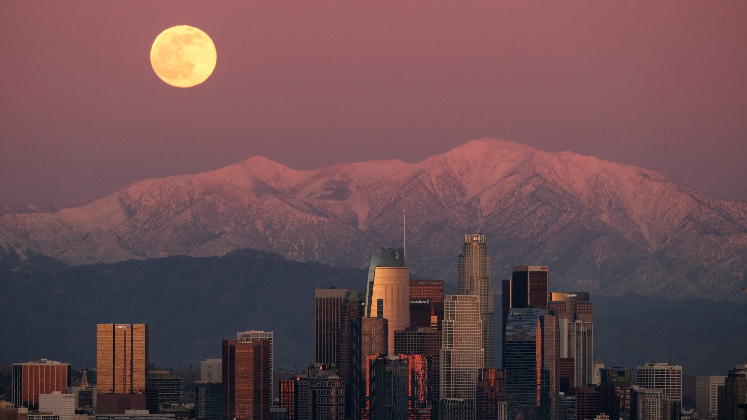 The moon rises over snow-covered mountains, behind the downtown Los Angeles skyline as seen from Kenneth Hahn State Recreation Area Tuesday, Dec. 29, 2020, in Los Angeles. (AP Photo/Ringo H.W. Chiu)