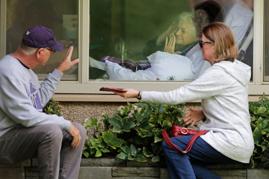 Judie Shape, center, who has tested positive for the coronavirus, blows a kiss to her son-in-law, Michael Spencer, left, as Shape's daughter, Lori Spencer, right, looks on, as they visit on the phone and look at each other through a window at the Life Care Center in Kirkland, Wash., on March 11, 2020. (Ted S. Warren / Associated Press)