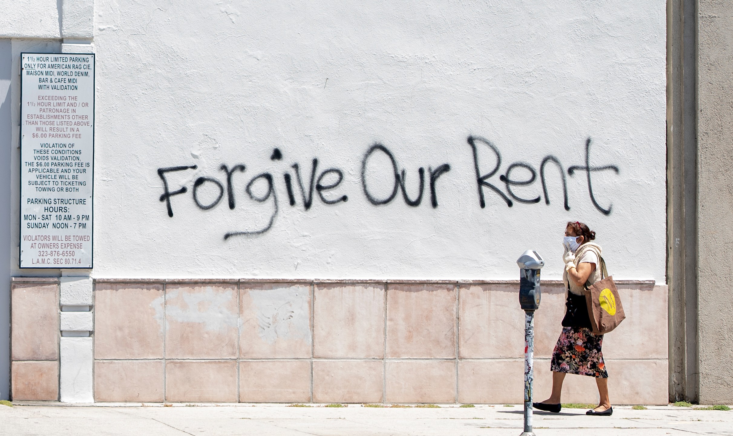 A woman wearing a mask walks past a wall bearing a graffiti asking for rent forgiveness on La Brea Ave on National May Day amid the Covid-19 pandemic, May 1, 2020, in Los Angeles, California. (Photo by VALERIE MACON/AFP via Getty Images)