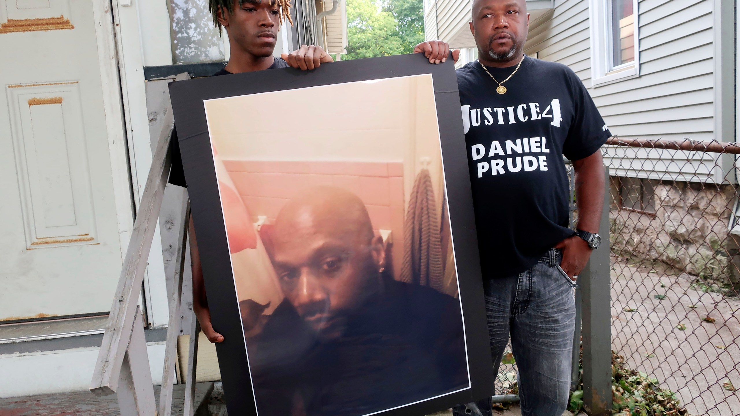 FILE — In this Sept. 3, 2020 file photo, Joe Prude, brother of Daniel Prude, right, and his son Armin, stand with a picture of Daniel Prude in Rochester, N.Y. Newly released transcripts show that a grand jury investigating the police suffocation death of Daniel Prude last year in Rochester, New York, voted 15-5 to clear the three officers involved in his restraint of a criminally negligent homicide charge sought by prosecutors. (AP Photo/Ted Shaffrey, File)