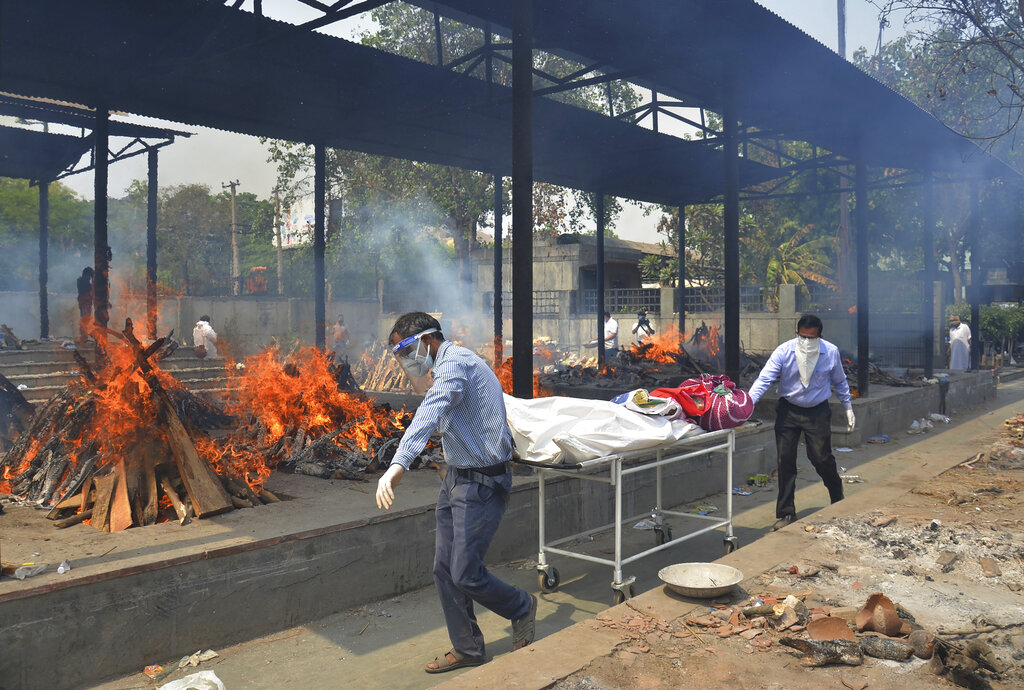 In this May 1, 2021, file photo, relatives carry the body of a person who died of COVID-19 as multiple pyres of other COVID-19 victims burn at a crematorium in New Delhi, India. (AP Photo/Amit Sharma, File)