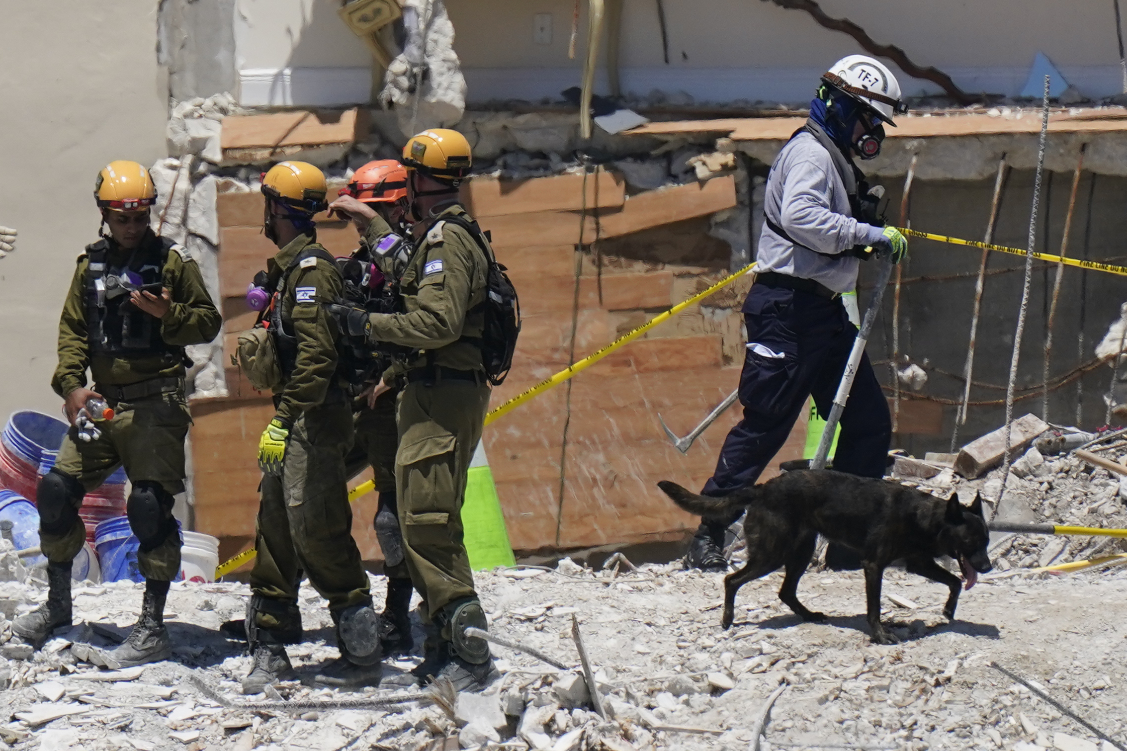 A dog aiding in the search walks past a team of Israeli search and rescue personnel, left, atop the rubble at the Champlain Towers South condo building, where scores of people remain missing one week after it partially collapsed, Friday, July 2, 2021, in Surfside, Fla. (AP Photo/Mark Humphrey)