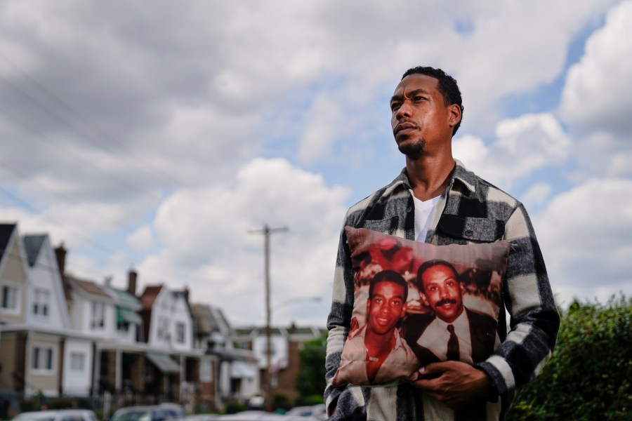 In this July 9, 2021, photo Brett Roman Williams poses for a photograph while holding a pillow with a photo of his father, Donald Williams, lower right, and brother Derrick Williams who both were killed by gunfire 20 years apart, in Philadelphia. (AP Photo/Matt Rourke)