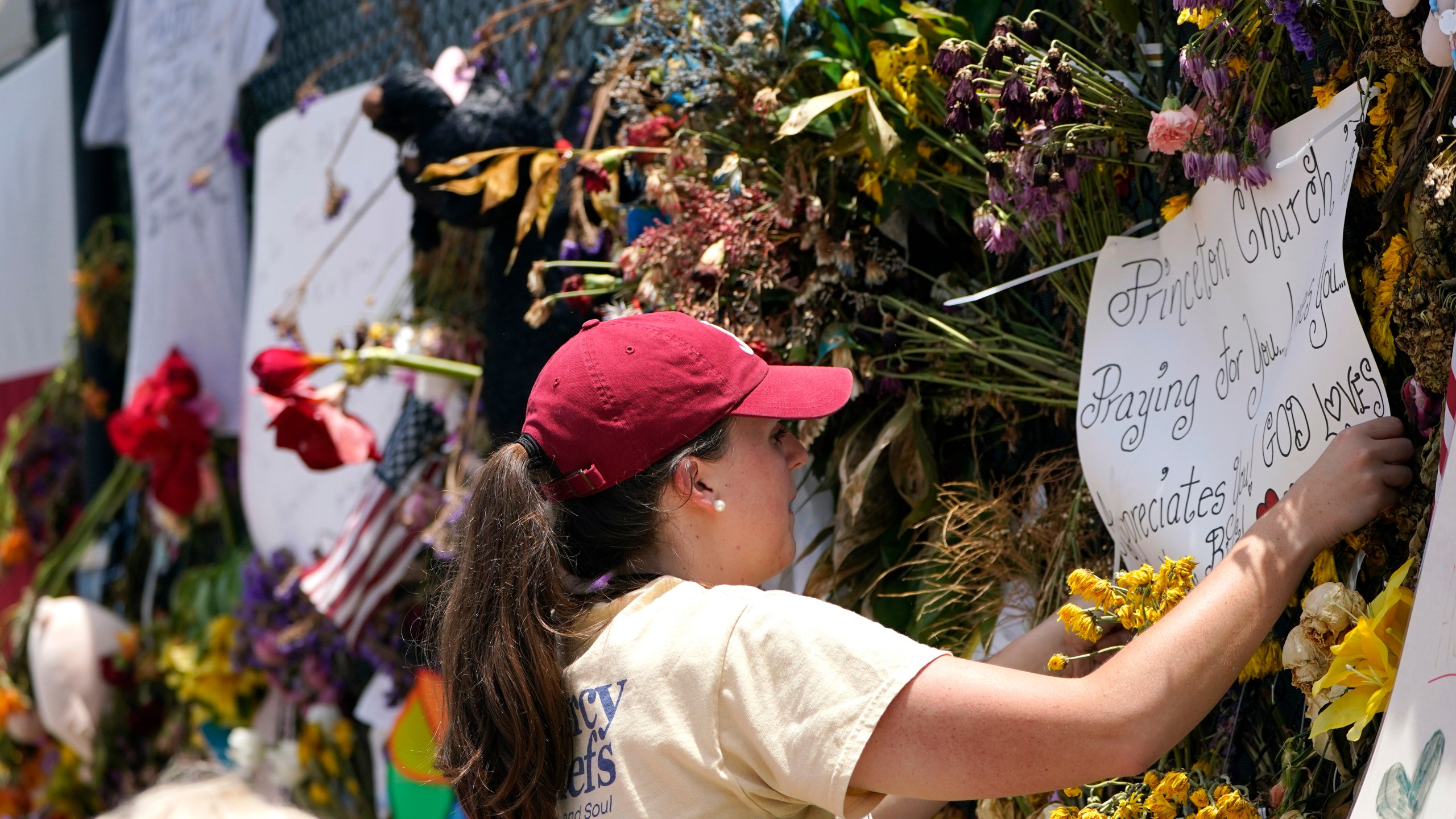 Molly MacDonald, with Mercy Chefs, hangs a sign on behalf of Princeton Church at a makeshift memorial remembering the victims of the nearby collapsed Champlain Towers South building, Wednesday, July 14, 2021, in Surfside, Fla. Mercy Chefs has set up a mobile kitchen to feed search and rescue teams working at the site three meals a day. (AP Photo/Lynne Sladky)