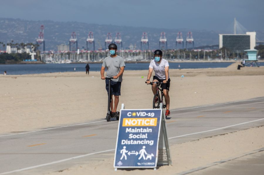 Men wearing facemasks ride a scooter and a bike near a notice about maintaining social distance on the beach in Long Beach on July 14, 2020. (APU GOMES/AFP via Getty Images)