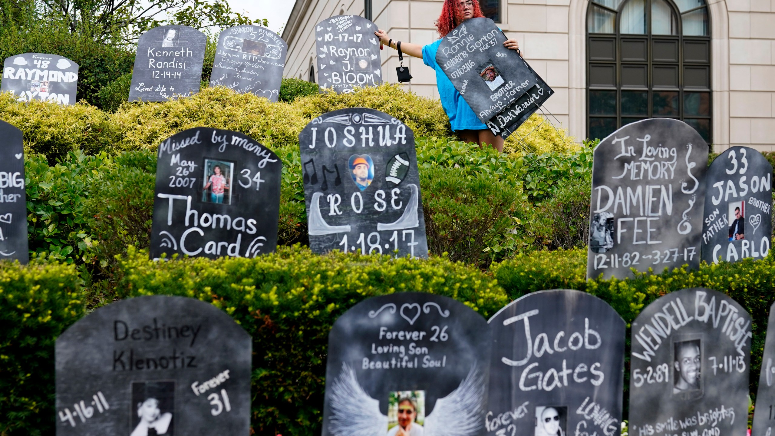 Jayde Newton helps to set up cardboard gravestones with the names of victims of opioid abuse outside the courthouse where the Purdue Pharma bankruptcy is taking place in White Plains, N.Y., Monday, Aug. 9, 2021. Purdue Pharma's quest to settle thousands of lawsuits over the toll of OxyContin is entering its final phase with the grudging acceptance of most of those with claims against the company. (AP Photo/Seth Wenig)