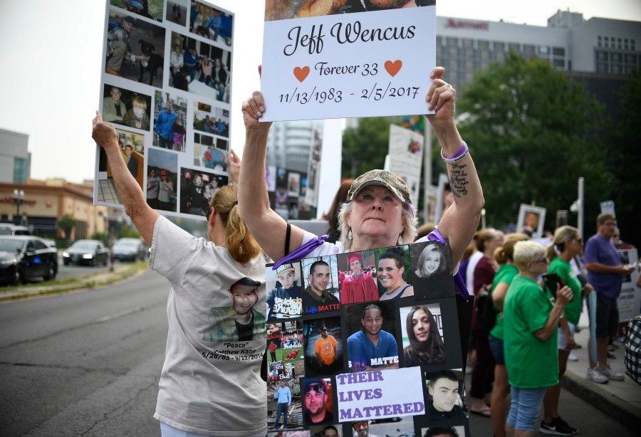 In this Aug. 17, 2018, file photo, Lynn Wencus of Wrentham, Mass., holds a sign with a picture of her son Jeff and wears a sign of others' loved ones lost to OxyContin and other opioids during a protest at Purdue Pharma LLP headquarters in Stamford, Conn. A landmark settlement in the nation’s opioid epidemic is forcing the owners of OxyContin maker Purdue Pharma to give up the company and pay out $4.5 billion. “Am I happy they don’t have to admit guilt and give up all their money? Of course not,” said Wencus. “But what would that do? It doesn’t bring my son back and it doesn’t help those who are suffering." (AP Photo/Jessica Hill, File)