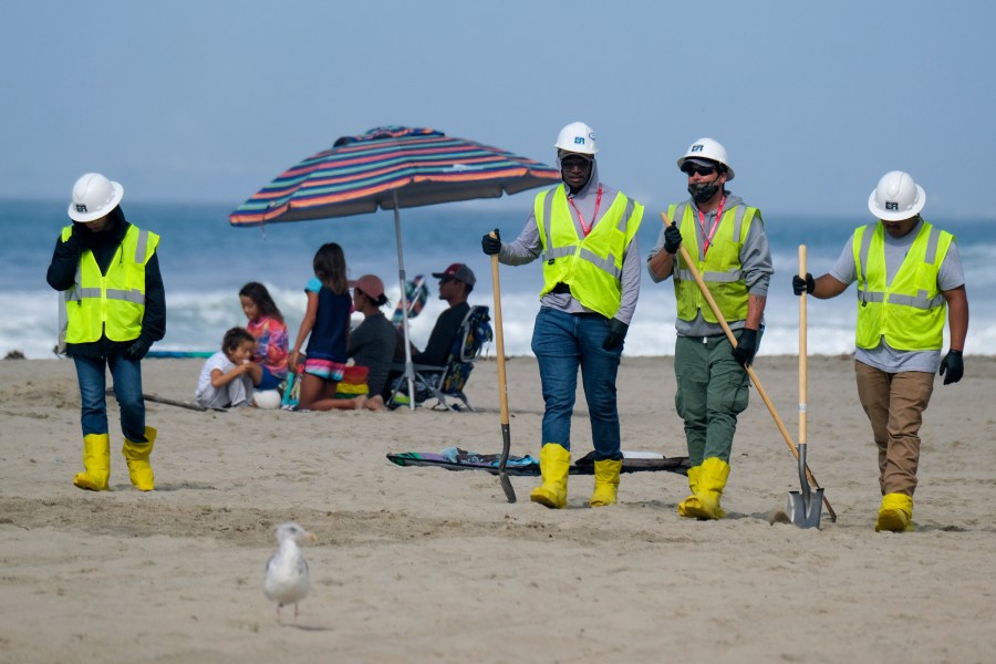 In this Oct. 11, 2021, file photo, a family, background, is under an umbrella as workers continue to clean the contaminated beach in Huntington Beach, Calif. California's uneasy relationship with the oil industry is being tested again by the latest spill to foul beaches and kill birds and fish off Orange County. (AP Photo/Ringo H.W. Chiu)