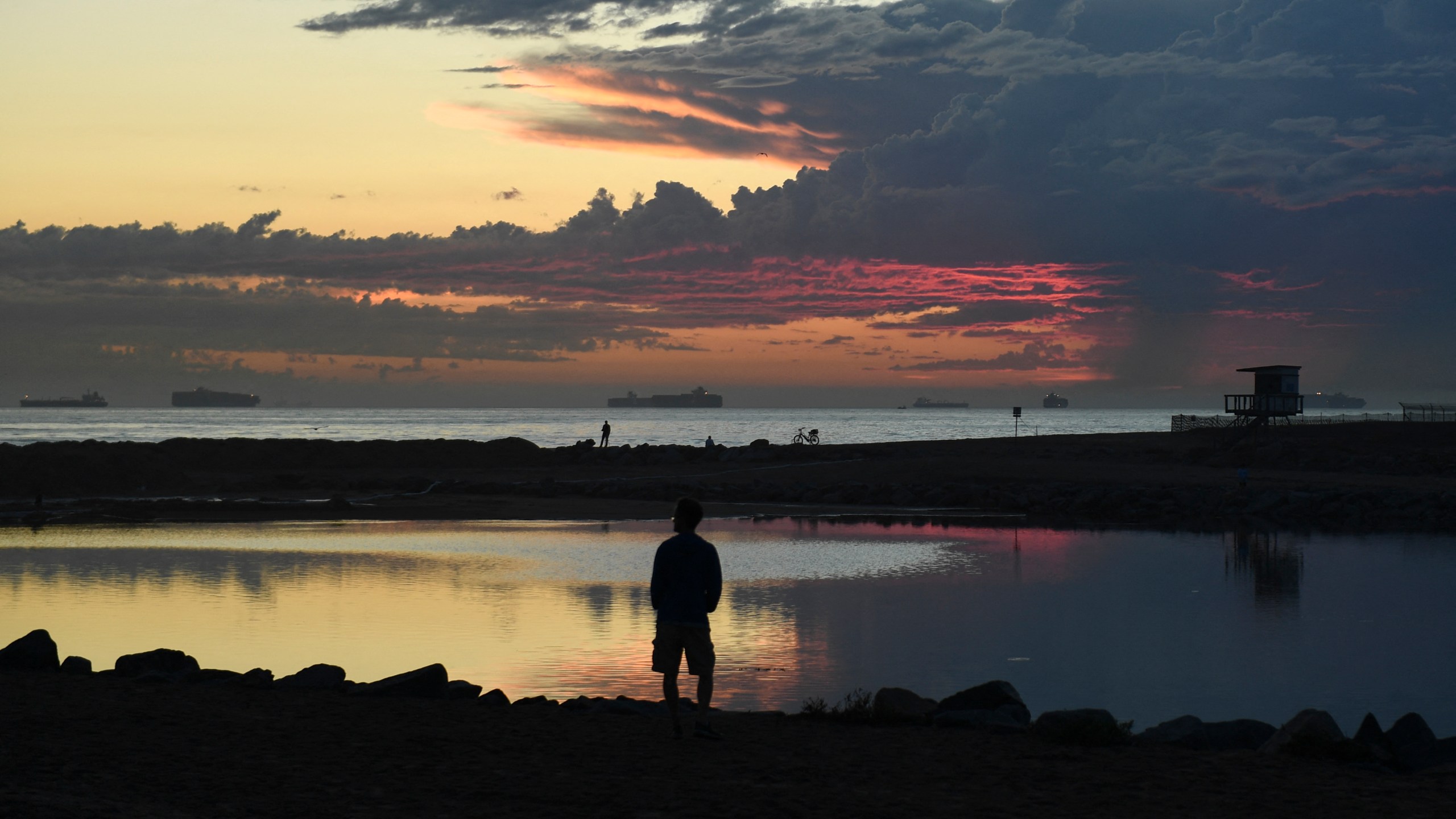 People watch the sunset from an inlet to the Santa Ana River blocked with sand and barriers to protect the inflow of oil as cargo container ships wait off the coast to enter the Port of Long Beach and Port of Los Angeles after an oil spill in the Pacific Ocean as seen from Huntington Beach, California on Oct. 4, 2021. (PATRICK T. FALLON/AFP via Getty Images)