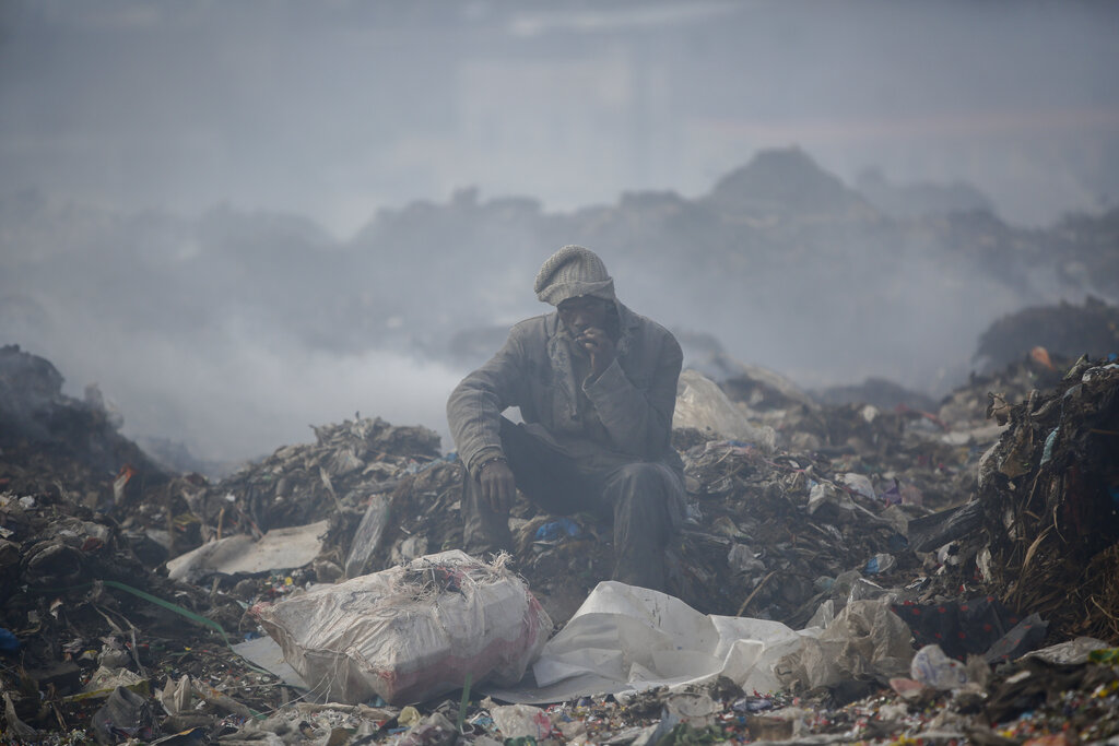 A man who scavenges recyclable materials for a living rests to smoke a cigarette on a mountain of garage amidst smoke from burning trash at Dandora, the largest garbage dump in the capital Nairobi, Kenya, Tuesday, Sept. 7, 2021. (AP Photo/Brian Inganga, File)