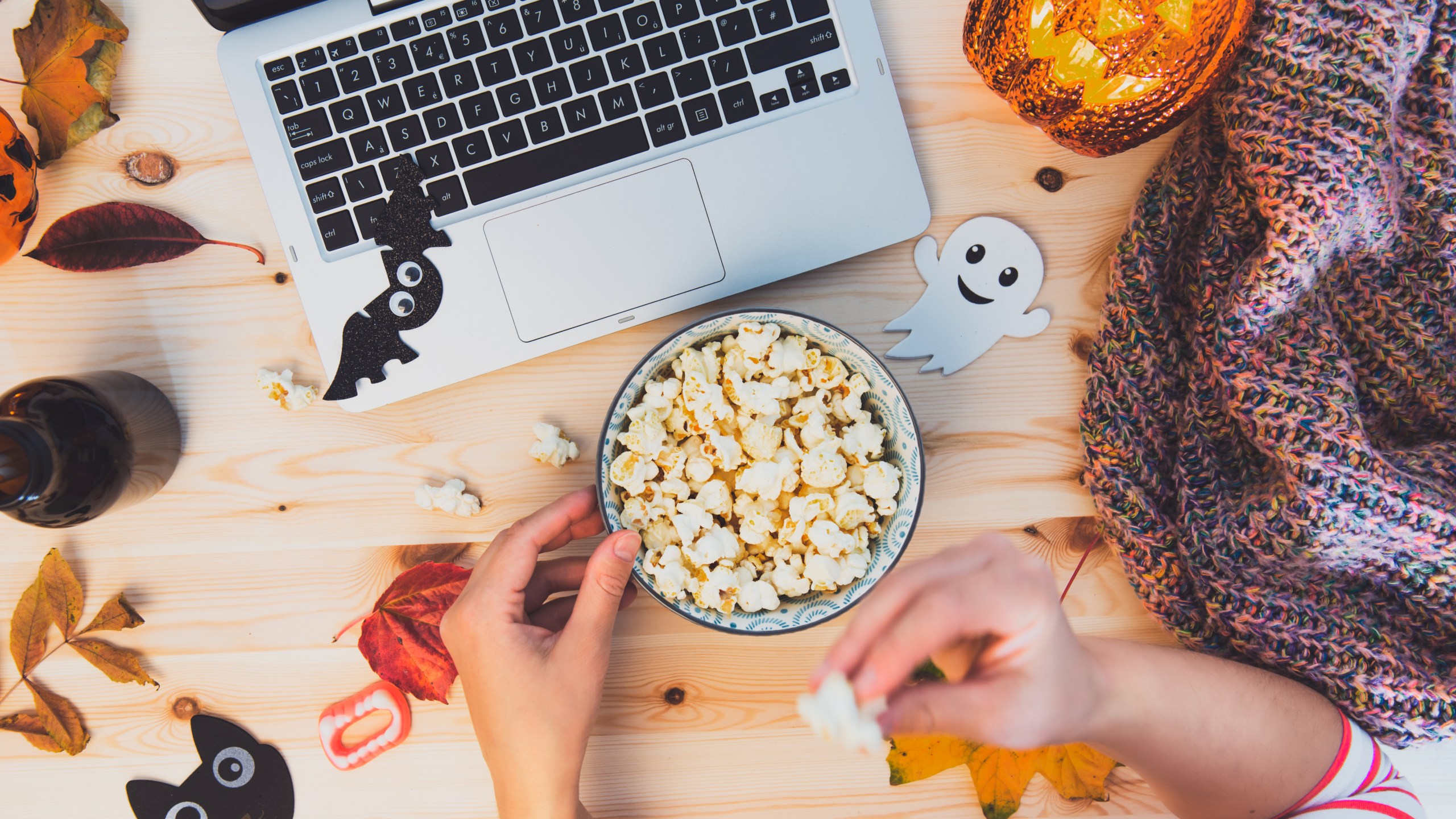 Woman eating popcorn while watching Halloween movie. Laptop, popcorn bowl, pumpkin, bats, ghost silhouettes, sweets, fall leaves, warm plaid on wooden background. Cozy and safe holiday party.