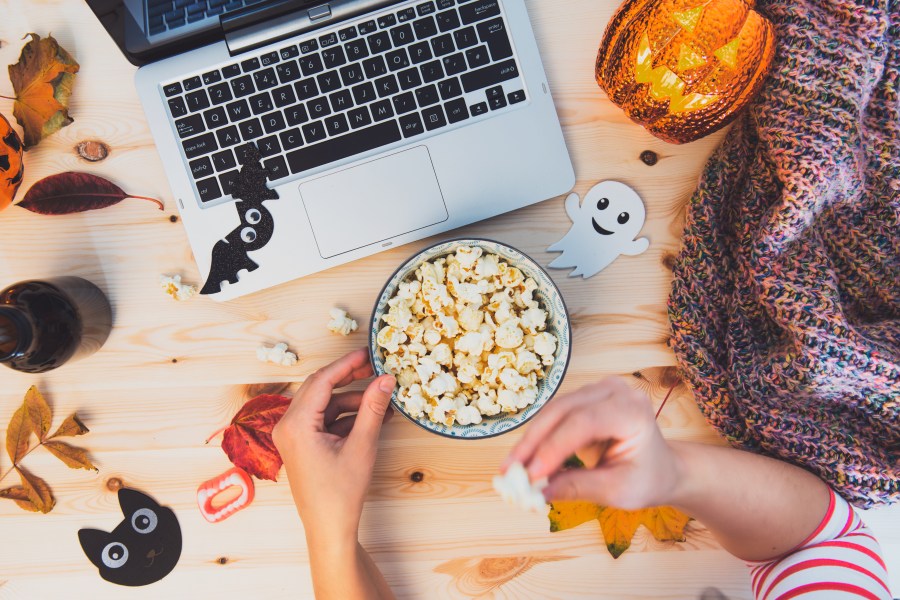 Woman eating popcorn while watching Halloween movie. Laptop, popcorn bowl, pumpkin, bats, ghost silhouettes, sweets, fall leaves, warm plaid on wooden background. Cozy and safe holiday party.