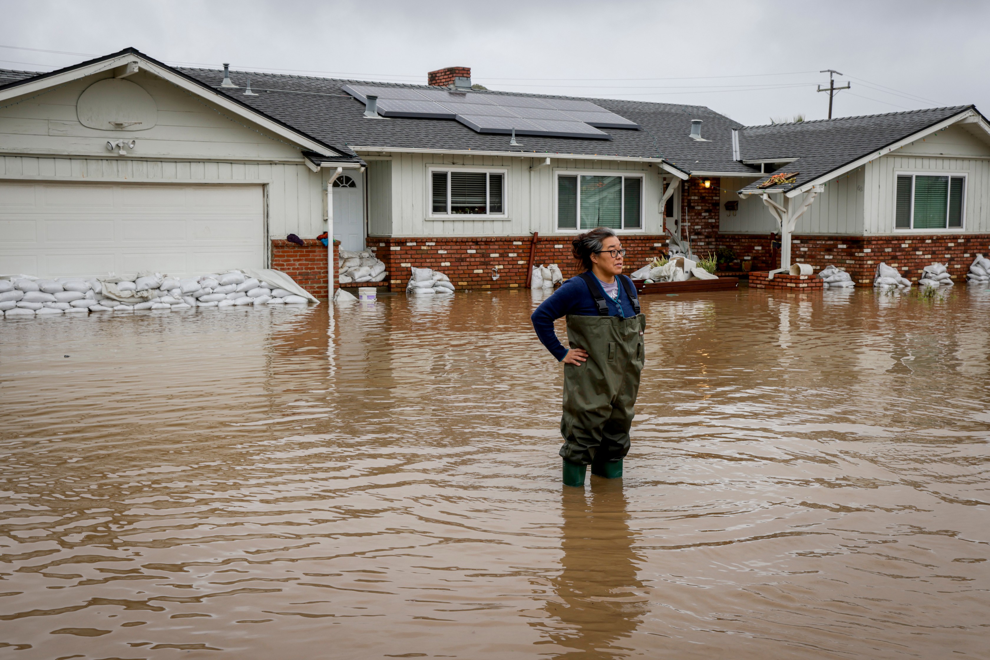 Colleen Kumada-McGowan stands in flood waters from huge amounts of rain in front of her home in a neighborhood off of Holohan Road near Watsonville, Calif. on Jan. 9, 2023.(Brontë Wittpenn/San Francisco Chronicle via AP)