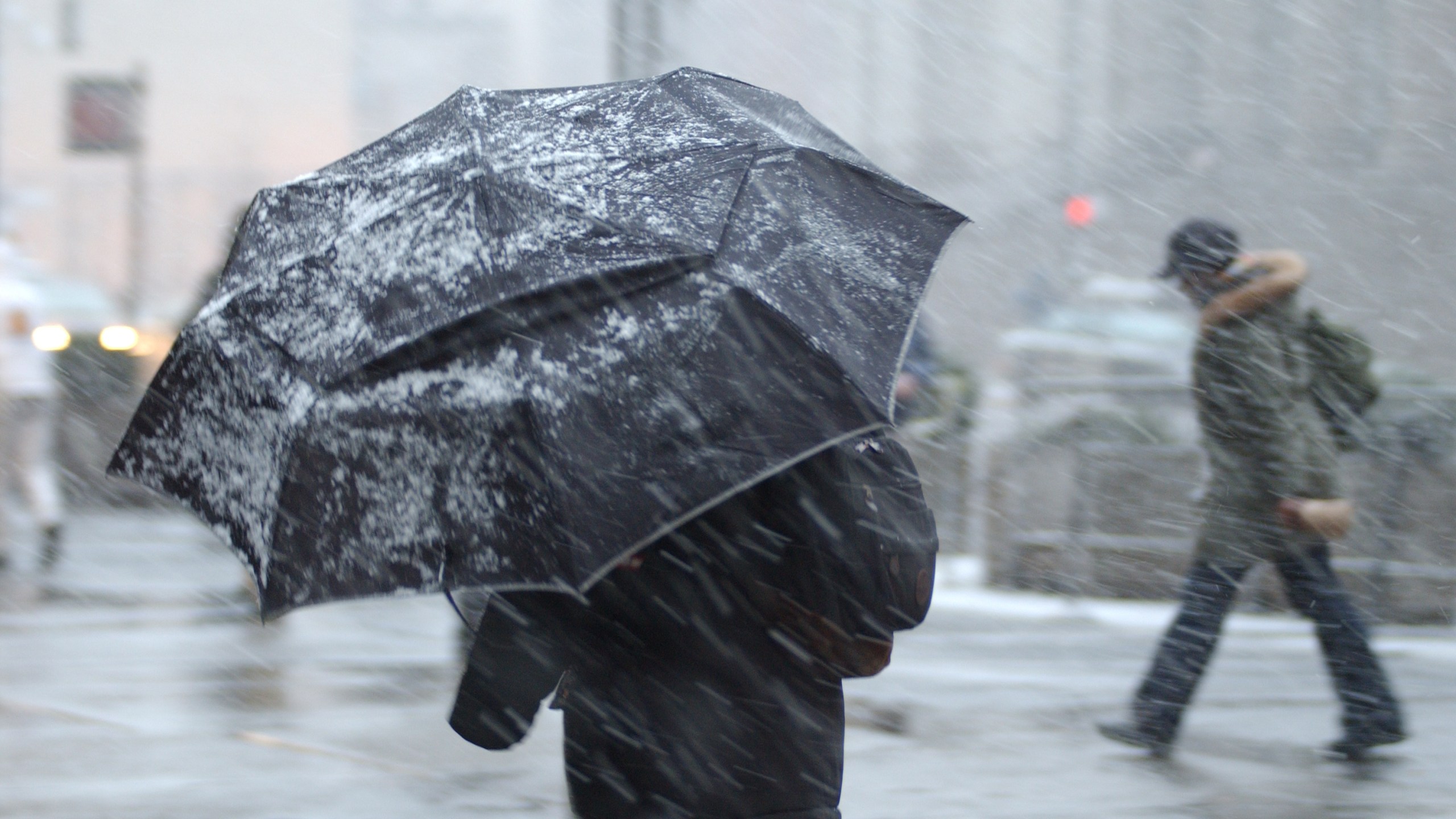 This is a photograph of a pedestrian in a snow storm with an aumbrella with a person in the background without an umbrella.