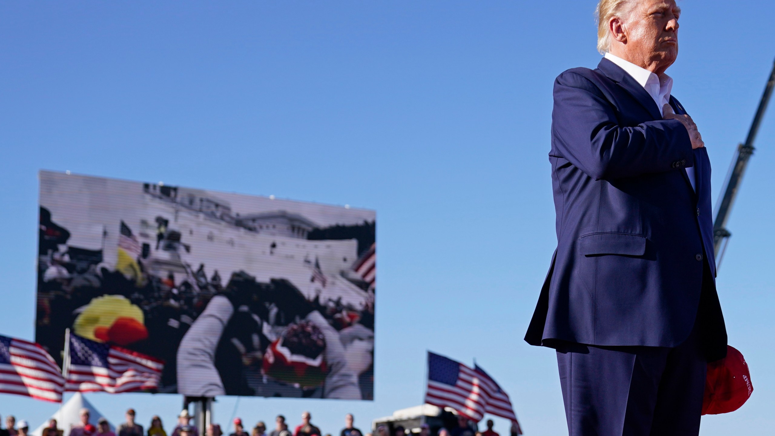 FILE - As footage from the Jan. 6, 2021, insurrection at the U.S. Capitol is displayed in the background, former President Donald Trump stands while a song, "Justice for All," is played during a campaign rally at Waco Regional Airport, Saturday, March 25, 2023, in Waco, Texas. The tune, “Justice for All,” is the Star-Spangled Banner and it was sung by a group of defendants jailed over their alleged roles in the January 2021 insurrection. The national anthem is overlaid with Trump reciting the Pledge of Allegiance. (AP Photo/Evan Vucci, File)