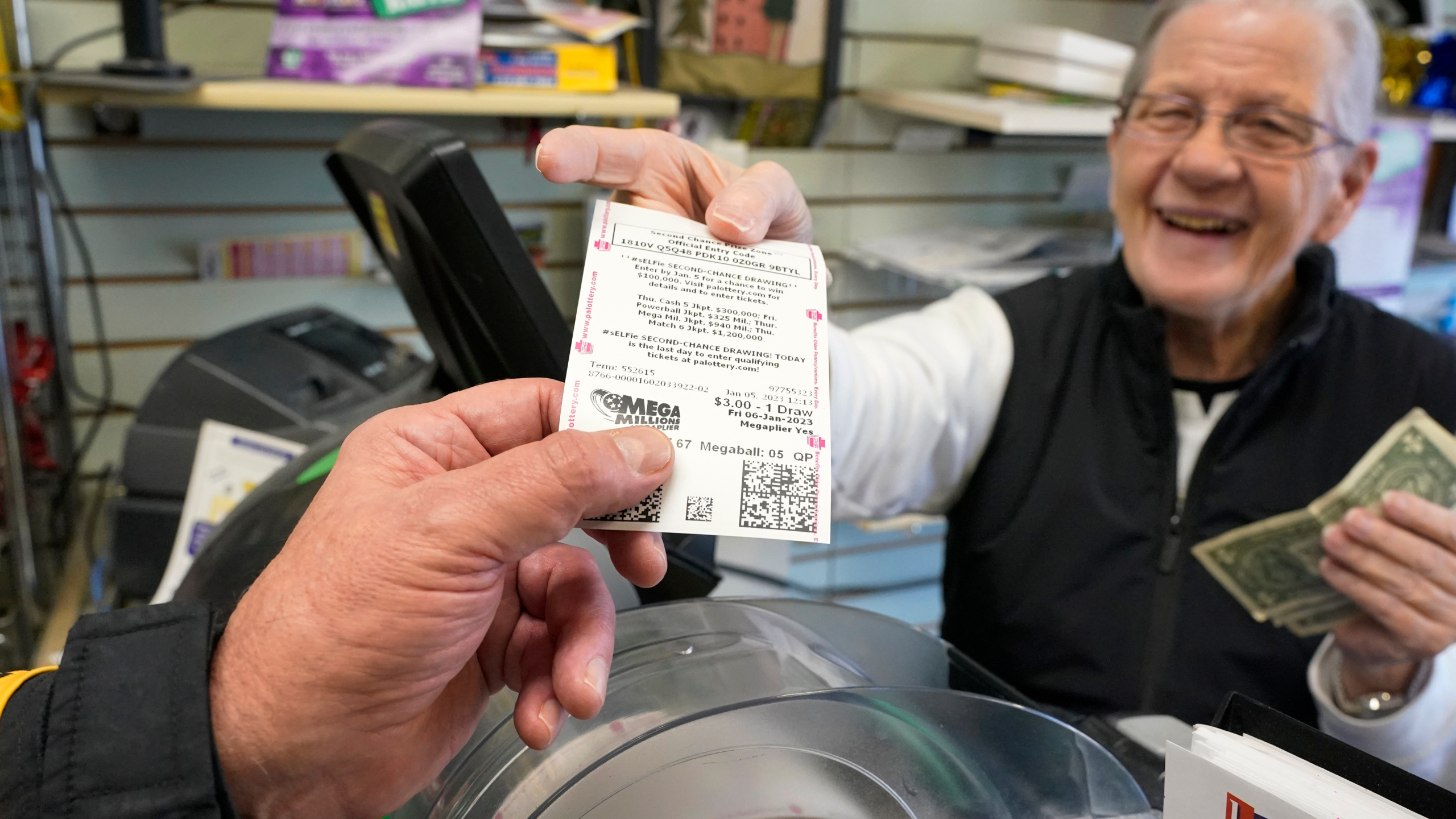 Dot Skoko, owner of Dot's Dollar More or Less shop in Mt. Lebanon, Pa., hands a customer a Mega Millions lottery ticket, Thursday, Jan. 5, 2022.