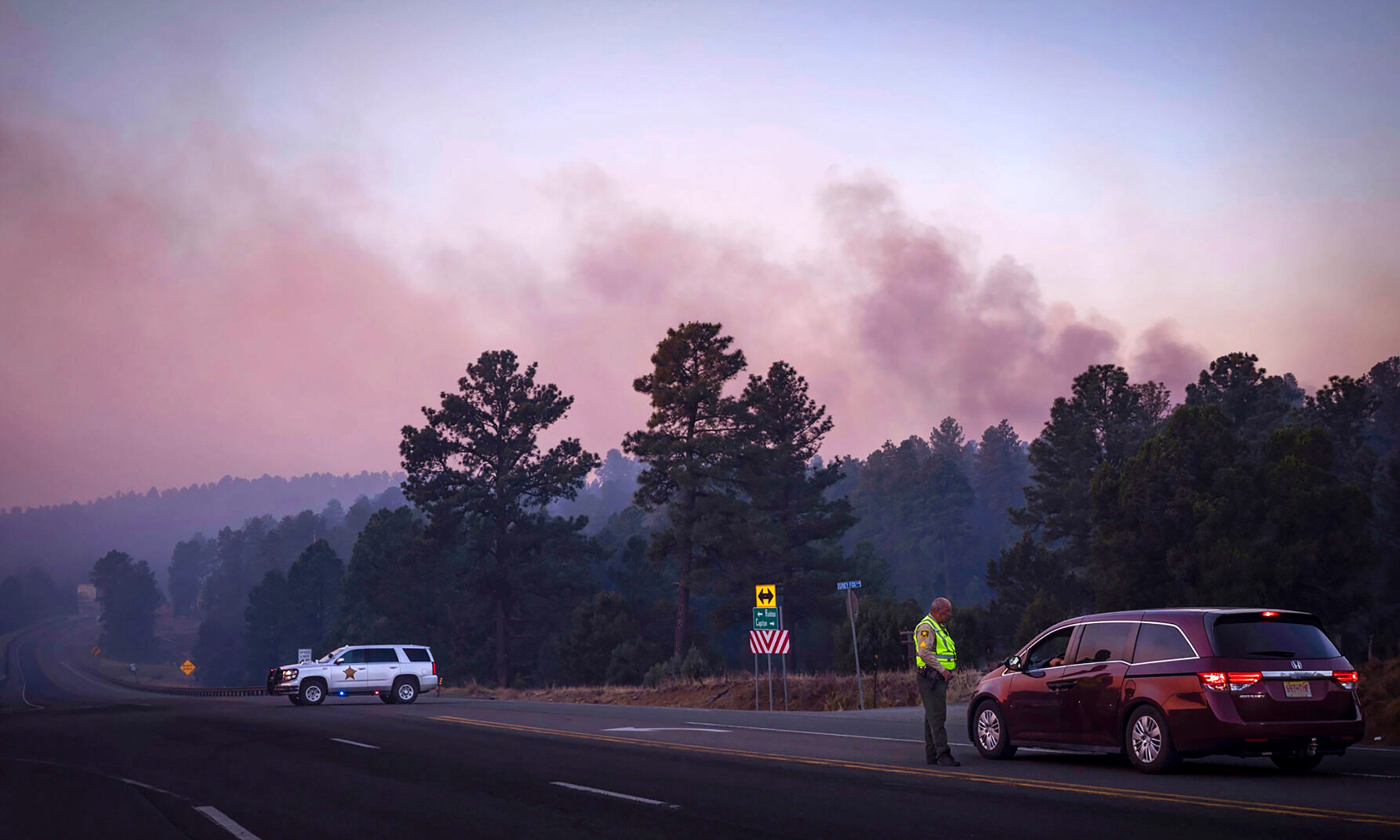 Smoke from the South Fork Fire reflects a pink hue during sunrise Tuesday, June 18, 2024 as law enforcement officers turn drivers away on State Road 48 at State 220 because of wildfires burning near Ruidoso, N.M. The South Fork Fire has consumed nearly 14,000 acres. (Chancey Bush/The Albuquerque Journal via AP)