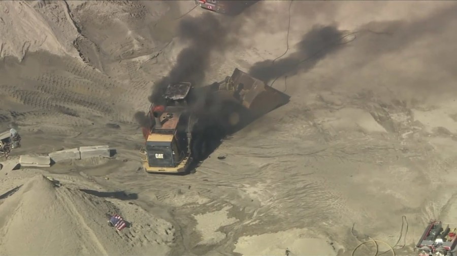 A burning articulated front loader is seen at a quarry after an explosion killed a Los Angeles County firefighter and injured another while they were battling a fire near Palmdale on June 14, 2024. (KTLA)