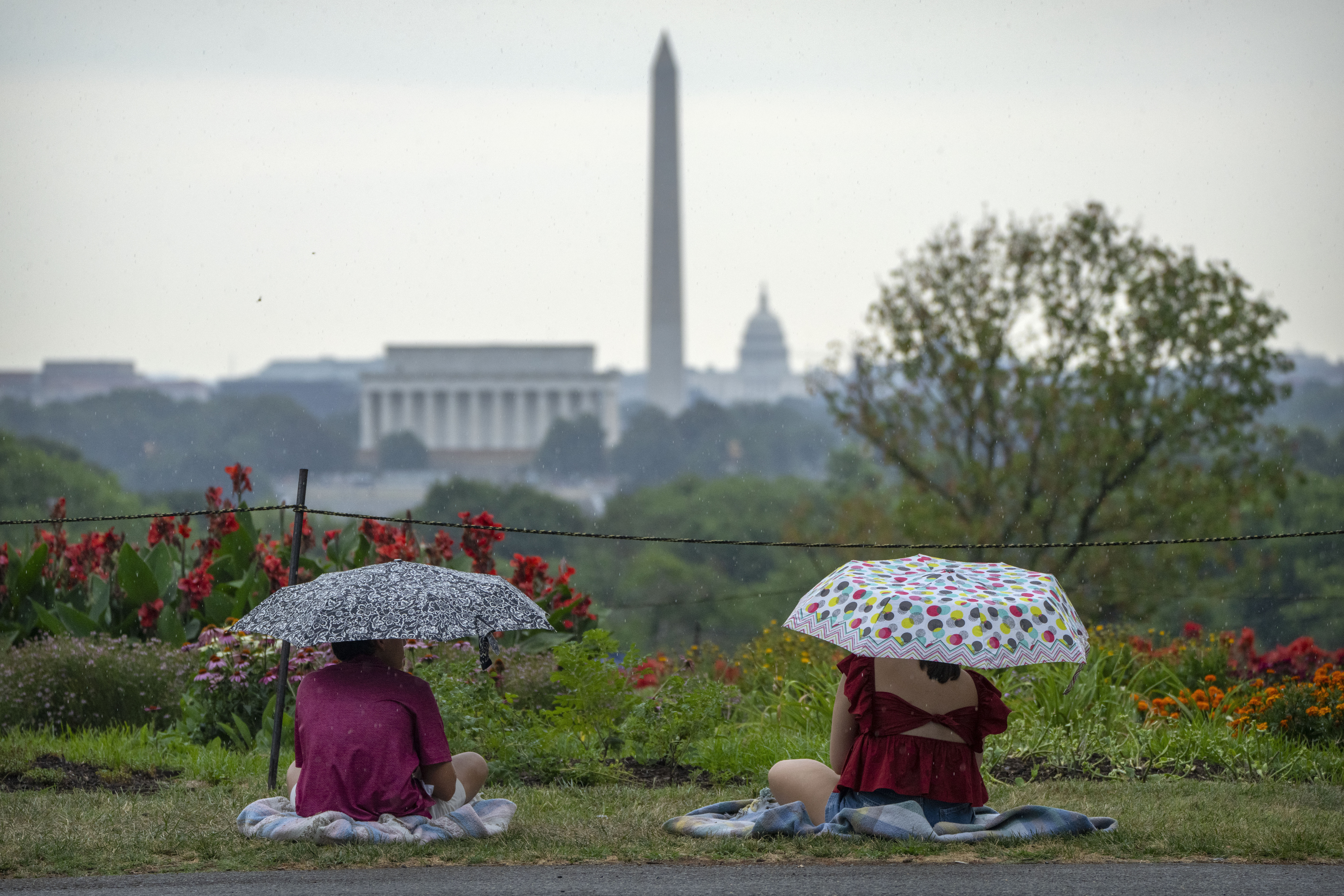People sit under umbrellas during a rain shower at the Netherlands Carillon on Thursday, July 4, 2024 in Arlington, Va., ahead of a fireworks display at the National Mall in Washington. (AP Photo/Mark Schiefelbein)
