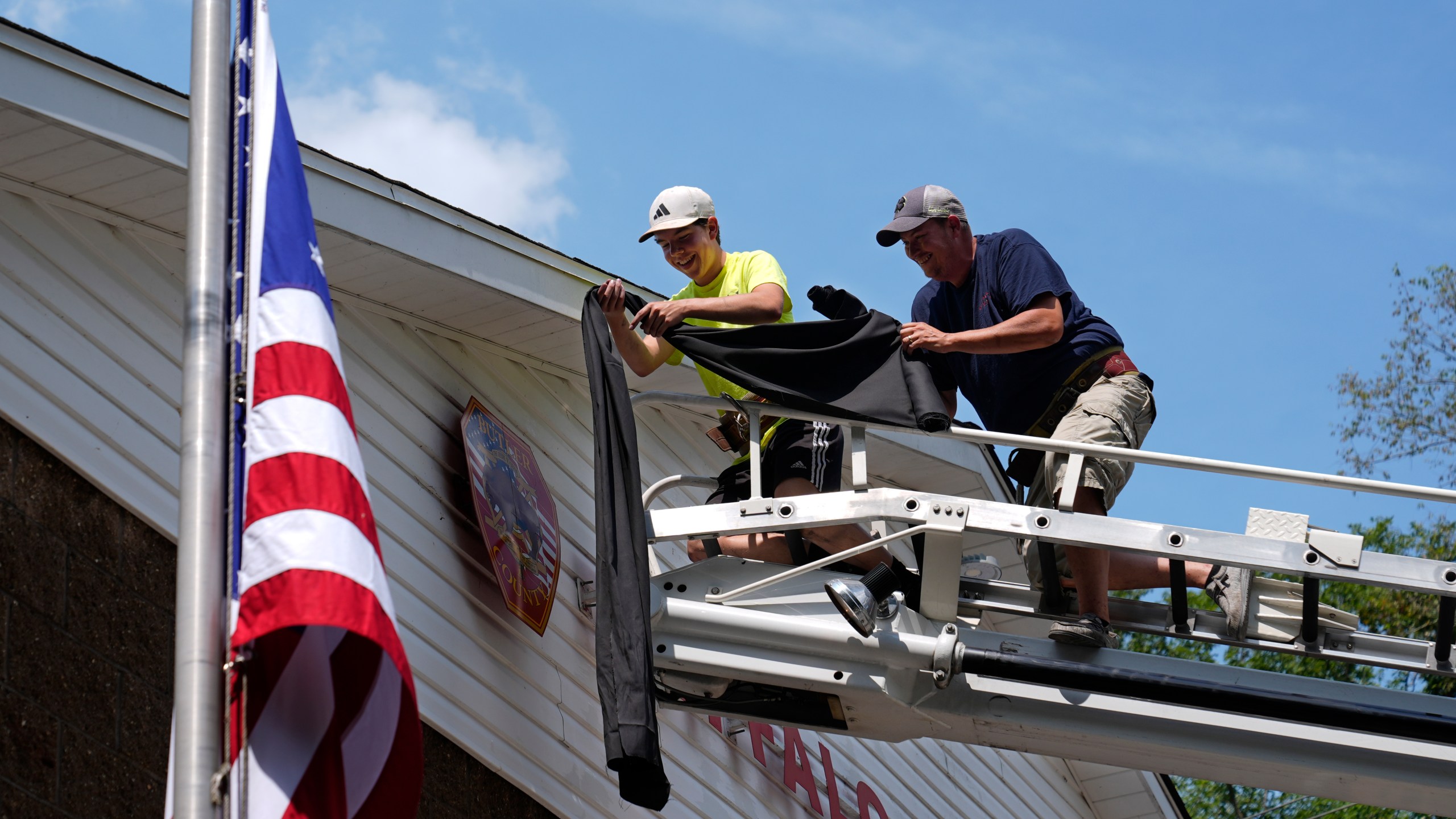 Logan Check, left, junior firefighter, and Randy Reamer, right, president and rescue captain at the Buffalo Township Fire Company 27, hang bunting on the fire station in memory of fellow firefighter Corey Comperatore, in Buffalo Township, Pa., Sunday, July 14, 2024. Comperatore was killed during a shooting at a campaign rally for Republican presidential candidate former President Donald Trump in Butler, Pa., on Saturday. The flag at the station house flies at half staff at left. (AP Photo/Sue Ogrocki)