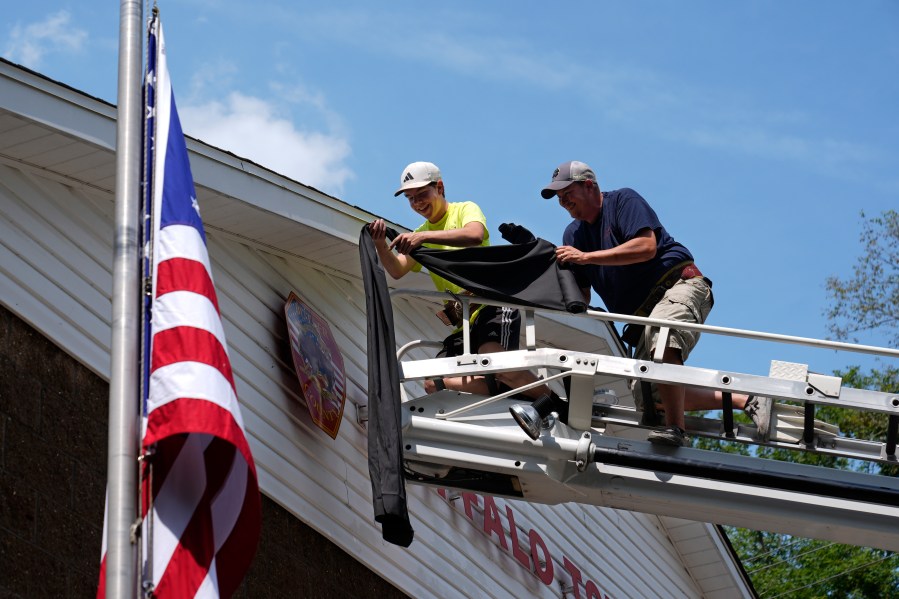 Logan Check, left, junior firefighter, and Randy Reamer, right, president and rescue captain at the Buffalo Township Fire Company 27, hang bunting on the fire station in memory of fellow firefighter Corey Comperatore, in Buffalo Township, Pa., Sunday, July 14, 2024. Comperatore was killed during a shooting at a campaign rally for Republican presidential candidate former President Donald Trump in Butler, Pa., on Saturday. The flag at the station house flies at half staff at left. (AP Photo/Sue Ogrocki)