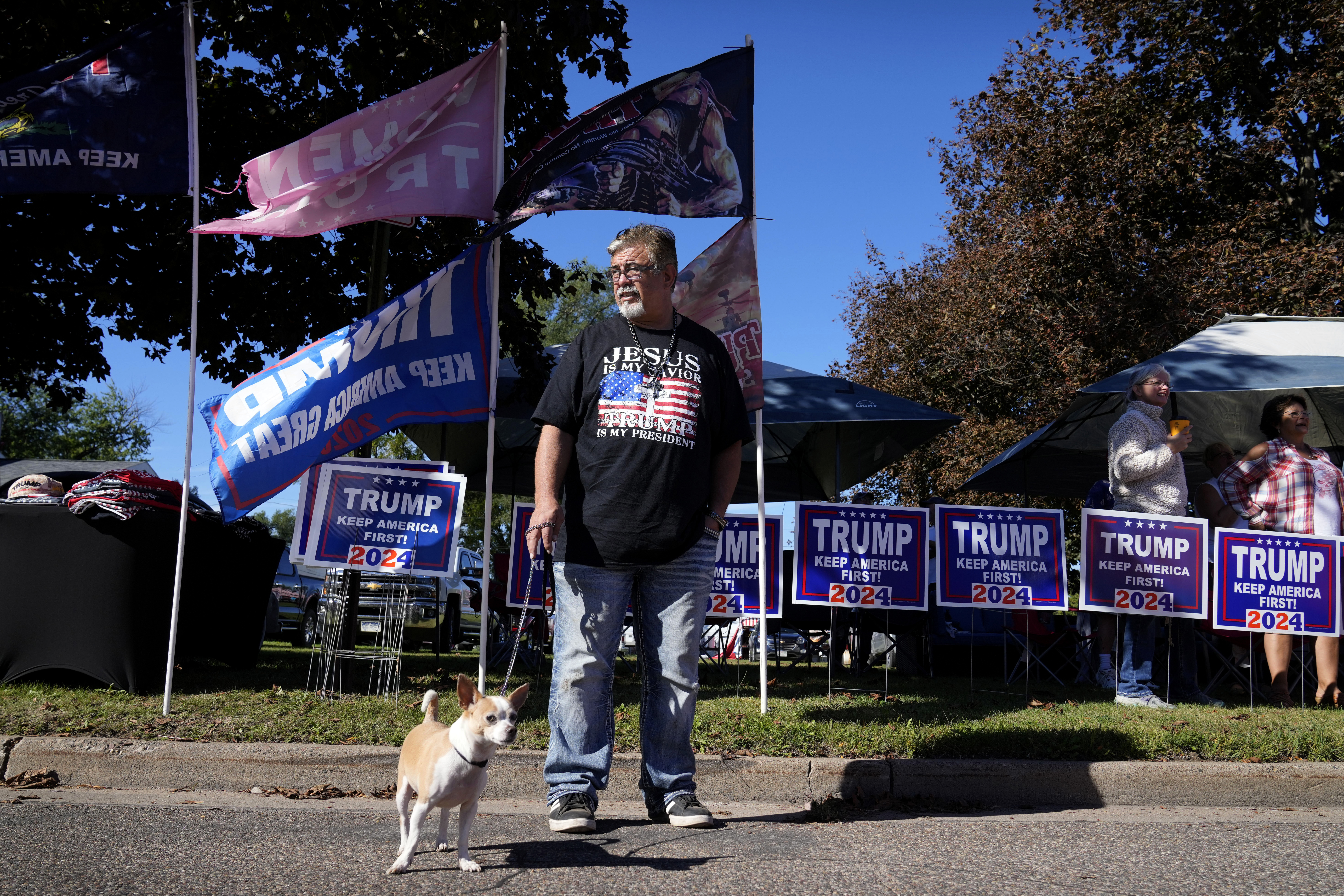 Merchandise vendor Sam Smith, of Sarasota, Fla., stands with his dog Milo while waiting for Republican presidential nominee former President Donald Trump to arrive at a rally, Saturday, Sept. 28, 2024, in Prairie du Chien, Wis. (AP Photo/Charlie Neibergall)