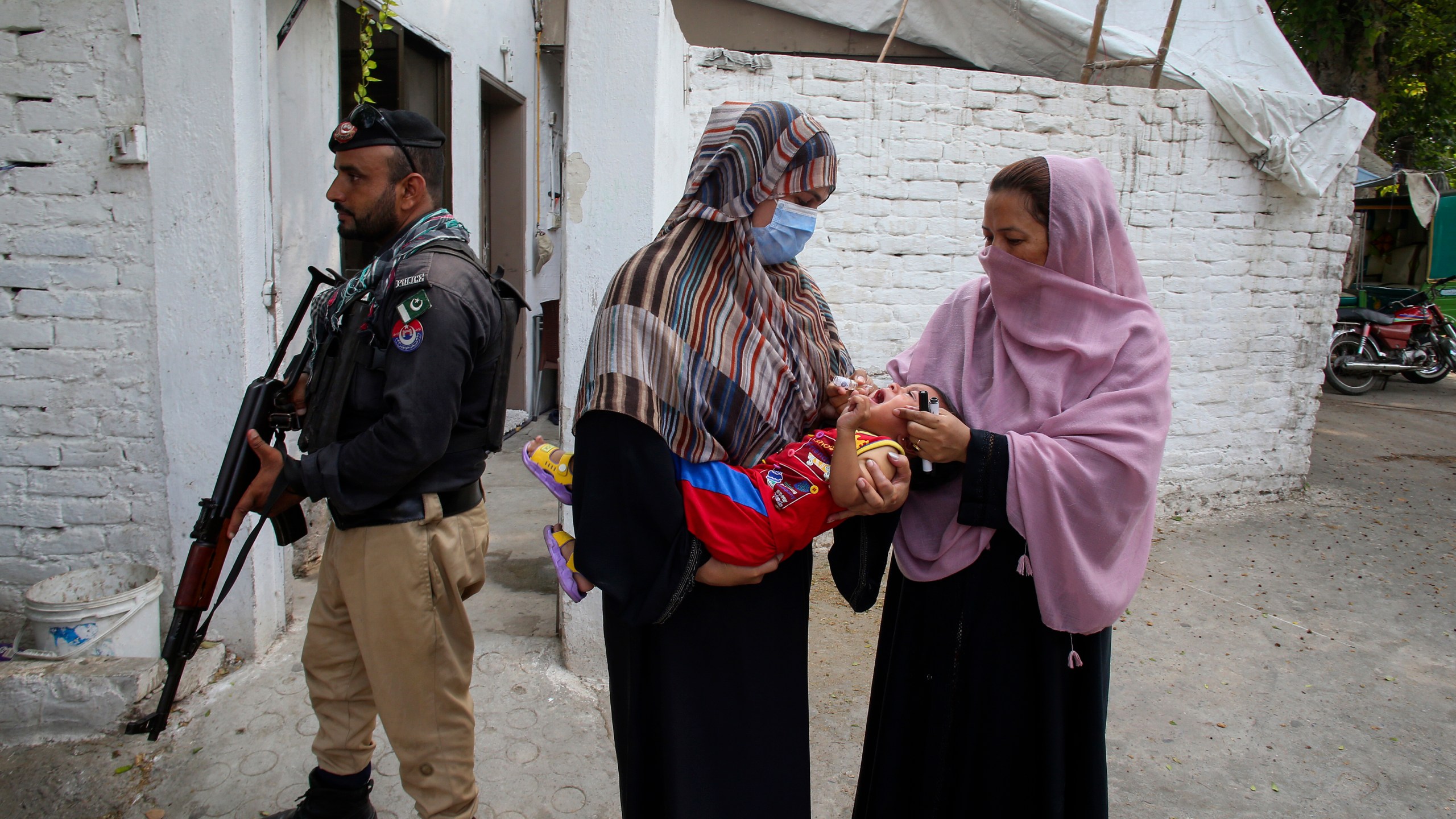 FILE -A police officer stands guard as a health worker, right, administers a polio vaccine to a child in a neighborhood of Peshawar, Pakistan, Sept. 9, 2024. (AP Photo/Muhammad Sajjad, File)