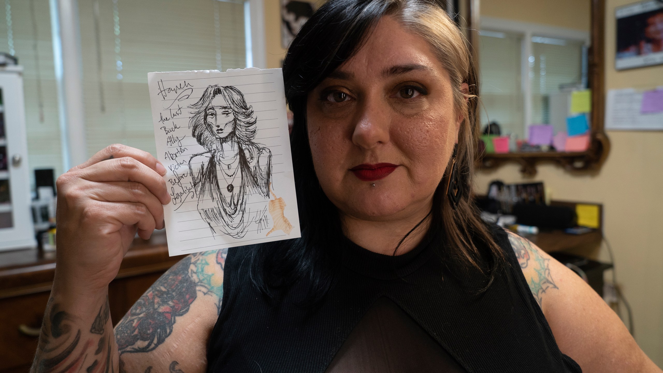 Tattoo artist Liz "Gruesome" Haycraft poses with a drawing of a fictitious back-alley abortion victim at her Breathless Arts Tattoo studio in Fayetteville, N.C., on Monday, Sept. 30, 2024. Haycraft drew it the day the U.S. Supreme Court overturned Roe v. Wade, which had previously legalized abortion nationwide. (AP Photo/Allen G. Breed)