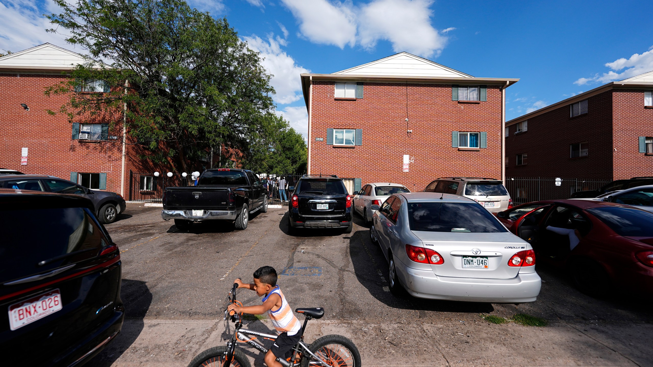 FILE - A boy rides his bicycle past apartment buildings as a rally staged by the East Colfax Community Collective is held in the courtyard to address chronic problems in the apartment buildings occupied by people displaced from their home countries in central and South America, Sept. 3, 2024, in Aurora, Colo. (AP Photo/David Zalubowski, File)