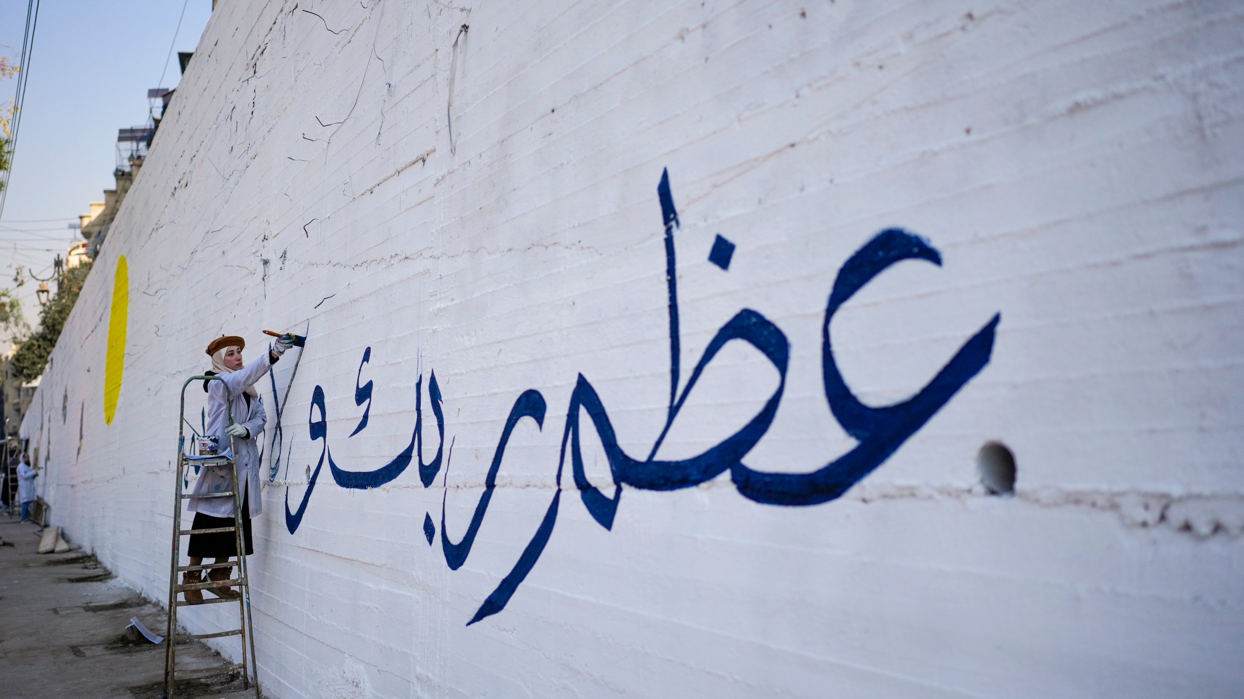 A girl writes "Worship your God" in Arabic as a group of young volunteers paints a mural symbolizing peace on a wall on the outskirts of Damascus, Syria, Sunday Jan. 12, 2025. (AP Photo/Mosa'ab Elshamy)